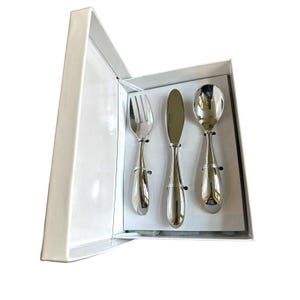May include: A silver-colored, three-piece flatware set, including a fork, knife, and spoon, presented in a white gift box. The utensils have a teardrop-shaped handle design. The set is ideal for a baby or toddler.