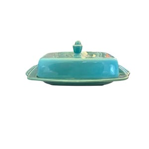 May include: A turquoise ceramic butter dish with a lid and a small handle. The dish has a rectangular shape with rounded edges and a scalloped design on the sides. The lid has a small, rounded handle.