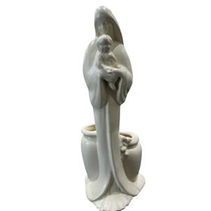 May include: A glossy off-white ceramic figurine depicting a tall, draped figure holding a small child. The base of the sculpture incorporates two hollow, rounded vessel-like forms on either side of the central figure's long, flowing robes.