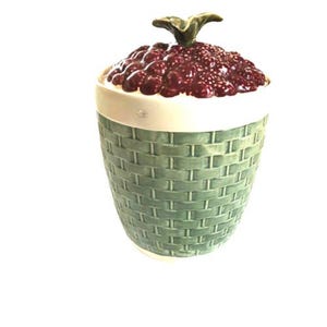 May include: A ceramic jar with a lid shaped like a cluster of dark red raspberries topped with a green stem. The body of the jar features a light green, embossed basket-weave pattern, while the upper rim is a smooth, solid white band.