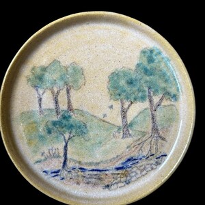 May include: A round ceramic plate with a hand-painted landscape scene. The design features green trees, a blue stream, and rolling hills in shades of green and blue. The plate has a beige rim and a speckled surface.