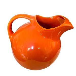 Op de afbeelding: Een glanzende, oranje keramische kan met een afgeronde body en een gebogen handvat. De kan heeft een tuit om gemakkelijk te schenken. De feloranje kleur geeft de kan een retro, mid-century modern gevoel.