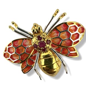 May include: A gold-toned bee brooch with red and orange enamel wings in a honeycomb pattern. The bee's body is gold, and the head features a cluster of small, red gemstones. The brooch has silver-colored legs and antennae.
