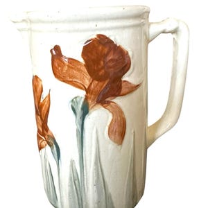 May include: Cream-colored ceramic pitcher with a handle and a spout. The pitcher is decorated with brown and orange iris flowers and green stems. The flowers are painted on the front of the pitcher.