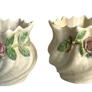 Puede incluir: Dos jarrones de cerámica color crema con borde festoneado y diseño en espiral. Cada jarrón está adornado con una rosa rosa, hojas verdes y pequeñas flores azules. Estos jarrones decorativos son ideales para plantas o flores pequeñas.