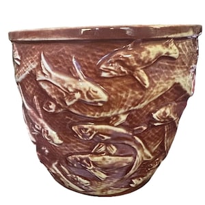 Puede incluir: Un cuenco de cerámica marrón con un relieve detallado de peces nadando en una red. Los peces son de un color beige claro, que contrasta con el marrón más oscuro del cuenco. El cuenco tiene un borde ligeramente ensanchado.