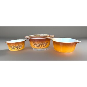 May include: A set of three glass kitchen bowls with a gradient orange and brown finish. The bowls feature a dark brown printed graphic of grapes and apples. The central bowl includes a matching glass lid with a clear handle and side grips.