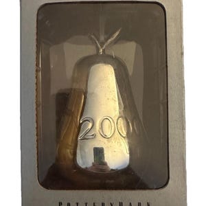 May include: A silver-colored pear-shaped ornament with the year 2000 embossed on the front. The ornament is housed in a rectangular cardboard box with a clear plastic window. The text "silver", "POTTERY BARN", and "2000 pear ornament" is printed.