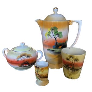 May include: A ceramic tea set featuring a tall lidded teapot, a lidded sugar bowl with two handles, a small pedestal cup, and a cylindrical mug. Each piece displays a hand-painted landscape with orange and yellow gradients, trees, and small houses.