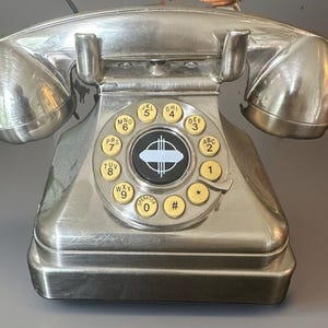 Puede incluir: Un teléfono de estilo giratorio en color plata metálico con una base rectangular y un auricular apoyado en la parte superior. El dial central incluye botones circulares de color crema con números y letras negros, rodeando una placa circular negra.
