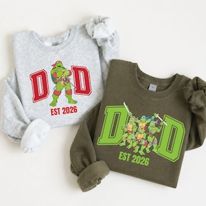 May include: A light gray crewneck sweatshirt featuring a red 'DAD' graphic with a Teenage Mutant Ninja Turtle and 'EST 2026' text, alongside an olive green crewneck sweatshirt with a green 'DAD' graphic, four turtles, and 'EST 2026' text.