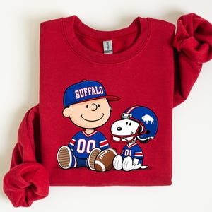 A red sweatshirt with a graphic of two cartoon characters, one wearing a football jersey and the other a baseball cap, sitting next to each other.