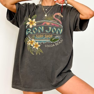 May include: A charcoal gray short-sleeve t-shirt featuring a graphic print with the text RON JON SURF SHOP COCOA BEACH. The design includes a pink flamingo, white and yellow tropical flowers, and green palm leaves in a vintage-inspired style.