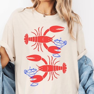 A woman wearing a white t-shirt with a graphic design of two red lobsters holding drinks.