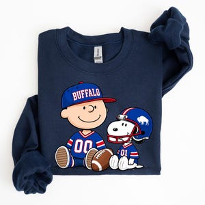 A navy blue sweatshirt with a graphic of two cartoon characters, one wearing a football jersey and the other a baseball cap, sitting together and smiling.