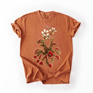May include: A burnt orange short-sleeve t-shirt featuring a central graphic illustration of a Venus flytrap plant with green and red leaves, white flowers, and a small blue dragonfly perched near the top of the botanical arrangement.