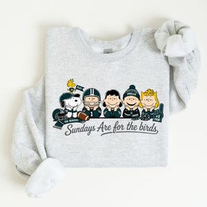 A gray sweatshirt with a graphic design featuring five cartoon characters, including a character holding a football, and the text "Sunday's Are for the birds" below the image.