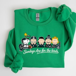 A green sweatshirt with a graphic design featuring five cartoon characters, including Snoopy, a character from Peanuts, and a football player. The text on the sweatshirt says "Sundays Are for the birds" and includes the phrase "GO WINS!"
