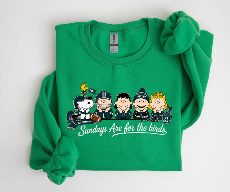 A green sweatshirt with a graphic design featuring five cartoon characters, including Snoopy, a character from Peanuts, and a football player. The text on the sweatshirt says "Sundays Are for the birds" and includes the phrase "GO WINS!"