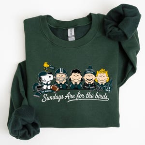 A green sweatshirt with a graphic design featuring five cartoon characters from the Peanuts comic strip, including Snoopy, Woodstock, Charlie Brown, Phil, and Bonnie. The text below the characters reads "Sundays Are for the birds".