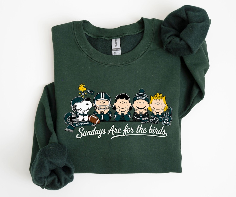 A green sweatshirt with a graphic design featuring five cartoon characters from the Peanuts comic strip, including Snoopy, Woodstock, Charlie Brown, Phil, and Bonnie. The text below the characters reads "Sundays Are for the birds".
