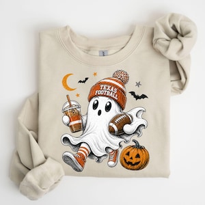 May include: A beige crewneck sweatshirt featuring a graphic of a ghost wearing an orange and white beanie with the text TEXAS FOOTBALL. The ghost holds an iced coffee and a football, with a jack-o'-lantern, bats, and a crescent moon nearby.