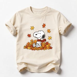 May include: A cream-colored short-sleeve t-shirt featuring a graphic of the cartoon character Snoopy wearing a red and black plaid scarf. Snoopy is sitting in a pile of orange, yellow, and brown autumn leaves with more leaves falling around.
