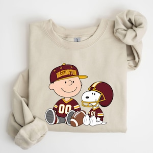 May include: A beige crewneck sweatshirt featuring a graphic of a cartoon character wearing a burgundy and gold baseball cap with the text WASHINGTON, alongside a cartoon dog in a football helmet, both wearing matching burgundy and gold jerseys.