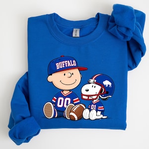A blue sweatshirt with a graphic of two cartoon characters, one wearing a football jersey and the other a baseball cap, sitting together and smiling.