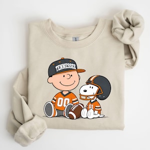 May include: A beige crewneck sweatshirt featuring a graphic of a cartoon character wearing a gray cap with the word TENNESSEE and an orange jersey, sitting next to a cartoon dog wearing a football helmet and an orange jersey with a football.