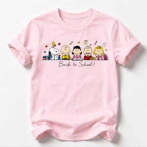 May include: A light pink short-sleeve t-shirt featuring a graphic of the Peanuts characters Snoopy, Woodstock, Charlie Brown, Lucy, Linus, and Sally resting their chins on their hands. The text Back to School! is printed below the characters.