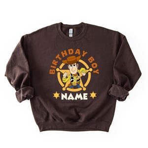 A brown sweatshirt with a cartoon character from the Toy Story franchise, specifically Woody, printed on it. The character is wearing a cowboy hat and holding a lasso. The sweatshirt has the text "BIRTHDAY BOY NAME" printed on it, indicating it is a birthday shirt for a boy named Woody.