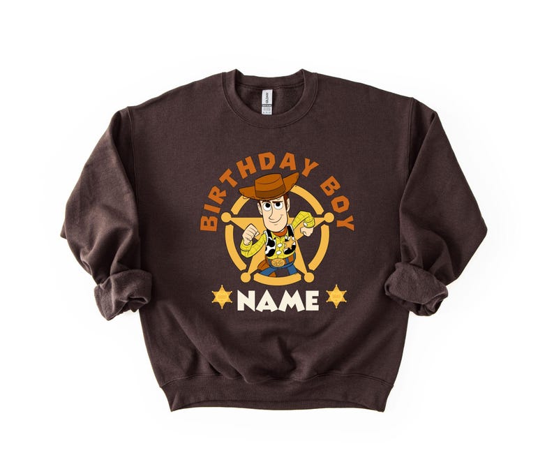A brown sweatshirt with a cartoon character from the Toy Story franchise, specifically Woody, printed on it. The character is wearing a cowboy hat and holding a lasso. The sweatshirt has the text "BIRTHDAY BOY NAME" printed on it, indicating it is a birthday shirt for a boy named Woody.
