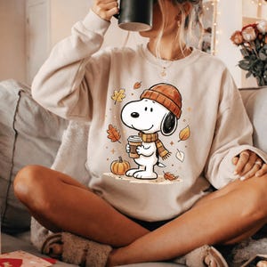 May include: A beige crewneck sweatshirt featuring a graphic of a cartoon dog wearing an orange beanie and a striped scarf, holding a coffee cup. The design includes falling autumn leaves and a small pumpkin, rendered in warm seasonal colors.