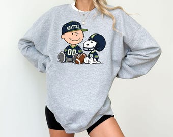 May include: A heather gray crewneck sweatshirt featuring a graphic print of two cartoon characters wearing dark blue and green football jerseys and helmets. One character wears a cap with the text SEATTLE, and both sit beside a brown football.