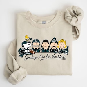 A beige t-shirt with a graphic design featuring five cartoon characters, including Snoopy, a football player, and other characters, along with the text "Sunday's Are for the birds".