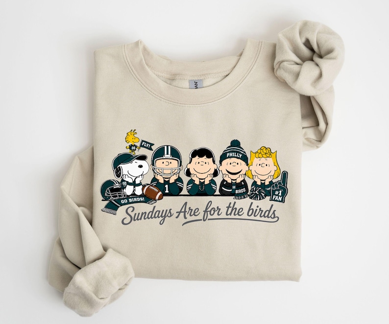 A beige t-shirt with a graphic design featuring five cartoon characters, including Snoopy, a football player, and other characters, along with the text "Sunday's Are for the birds".