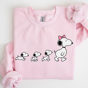 May include: A light pink sweatshirt featuring a Snoopy-themed graphic. The graphic shows Snoopy in four stages, with the final Snoopy wearing a pink bow. The sweatshirt has long sleeves and a crew neck.
