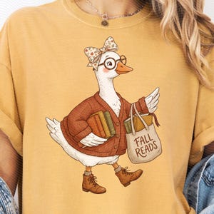May include: A mustard yellow t-shirt featuring a graphic of a white goose wearing a brown cardigan, round glasses, and a floral bow. The goose holds books and a tote bag with the text FALL READS printed on it, while wearing brown lace-up boots.