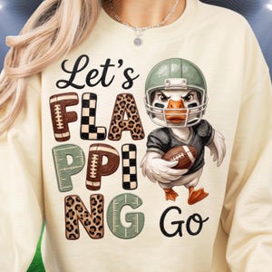 Puede incluir: Una sudadera color crema con cuello redondo que presenta un gráfico de un pato con casco y camiseta de fútbol americano, sosteniendo un balón. El texto Let's FLAPPING Go usa letras con patrones de fútbol, estampado de leopardo y cuadros.