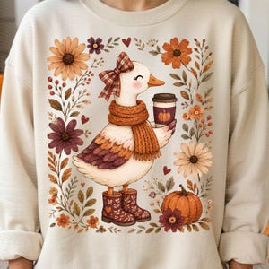 Può includere: Una felpa girocollo beige con la grafica di un'anatra bianca che indossa un fiocco a quadri, una sciarpa arancione a coste e stivali fantasia. L'anatra tiene in mano una tazza di caffè con una zucca stampata, circondata da fiori e foglie autunnali.