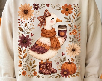 May include: A beige crewneck sweatshirt featuring a graphic of a white duck wearing a plaid bow, a ribbed orange scarf, and patterned boots. The duck holds a coffee cup with a pumpkin print, surrounded by autumn flowers, leaves, and a pumpkin.