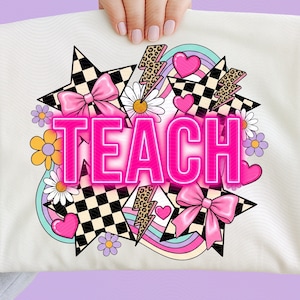 May include: A white t-shirt featuring a graphic design with the word TEACH in bright pink letters. The design includes black and white checkered stars, pink bows, leopard print lightning bolts, daisies, hearts, and pastel rainbow arches.