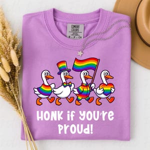 Puede incluir: Una camiseta lila Comfort Colors con un gráfico de cuatro gansos de dibujos animados con atuendos de arcoíris, marchando y sosteniendo una bandera de arcoíris. El texto "HONK if you're PROUD!" está impreso debajo.