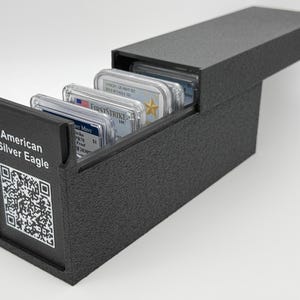 May include: A black textured storage box containing several rectangular coin slabs. The front panel features the text American Silver Eagle and a printed QR code. The box has a sliding lid design, with the interior compartments holding the coin cases.