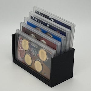 May include: A black coin holder containing several clear plastic cases. Each case holds multiple gold-colored coins. The cases are labeled with text, and the holder is on a white surface. The coins appear to be part of a collection.