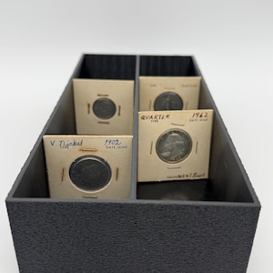 May include: A black, textured coin storage box with four compartments, each holding a coin in a cardboard holder. Two holders display the text "V Nickel" and "1902 DATE/MINT". The other two holders display the text "Quarter TYPE" and "1962 DATE/MINT".