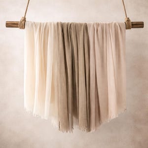 May include: Three scarves in cream, beige, and pale pink are draped over a wooden pole, suspended by rope. The scarves have a soft, flowing texture and a fringed edge. The neutral colours suggest a focus on natural fibres and minimalist design.