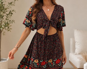 Puede incluir: Un vestido negro de manga corta con un estampado floral vibrante de flores rojas y amarillas. La prenda tiene un escote en V profundo con cierre de lazo frontal, un detalle de abertura en la cintura y una falda acampanada sobre la rodilla.