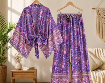 Puede incluir: Un conjunto coordinado morado con estampado floral, compuesto por un kimono y pantalones de pernera ancha. El kimono tiene un cierre de lazo y los pantalones tienen cintura con cordón. El conjunto presenta un patrón vibrante con acentos rosas, azules y amarillos sobre fondo morado.